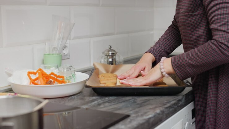 Signora che appoggia delicatamente biscotti appena sfornati su carta da forno, usando un cartone animato per asciugare l'olio in eccesso, circondata da utensili da cucina e ingredienti come formine per biscotti e guarnizioni colorate