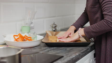 dama colocando suavemente galletas recién horneadas en papel de pergamino, usando cartón para secar el exceso de aceite, rodeada de utensilios de cocina e ingredientes como cortadores de galletas y ingredientes coloridos