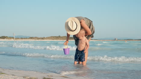 una madre joven hablando con su hijo en la orilla del mar. un bebé lindo caminando a lo largo del surf.