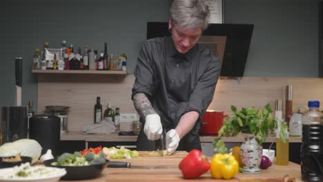 Young-professional-male-chef-in-an-elegant-black-shirt-with-an-alternative-look,-tattoos,-gloves-chopping-fresh-leek-A-modern,-fancy-looking-kitchen-with-fresh-vegetables-besides-Cinematic-Dolly-4K