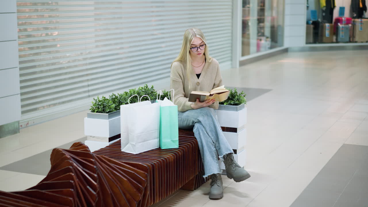 dama sentada en un centro comercial brillante con las piernas cruzadas, leyendo su libro, con bolsas de compras cerca, ambiente relajado, aspecto casual, con un centro comercial moderno en el fondo