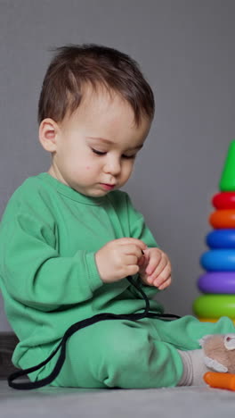 lovely kid sits on the floor holding a shoe lace and taking it to his mouth. toy are around the baby. indoors backdrop. vertical video