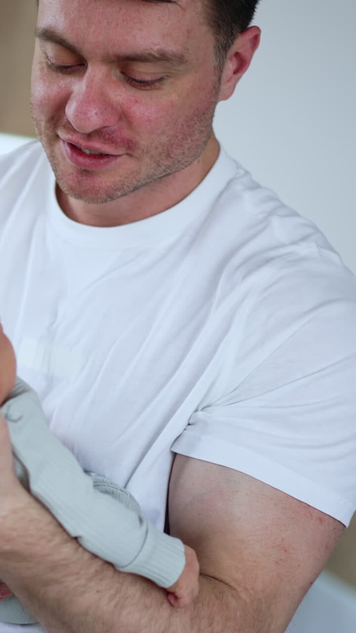 Broad-shouldered man wearing white t-shirt waving his newborn child. Dad taking care of the child while mom is resting. Vertical video
