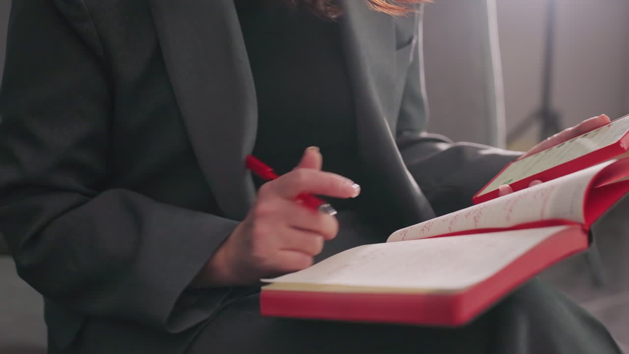 Businesswoman writing in red notebook with pen, focused on work, modern office environment, gray suit, sitting relaxed, productivity, decision-making, corporate workspace, concentration