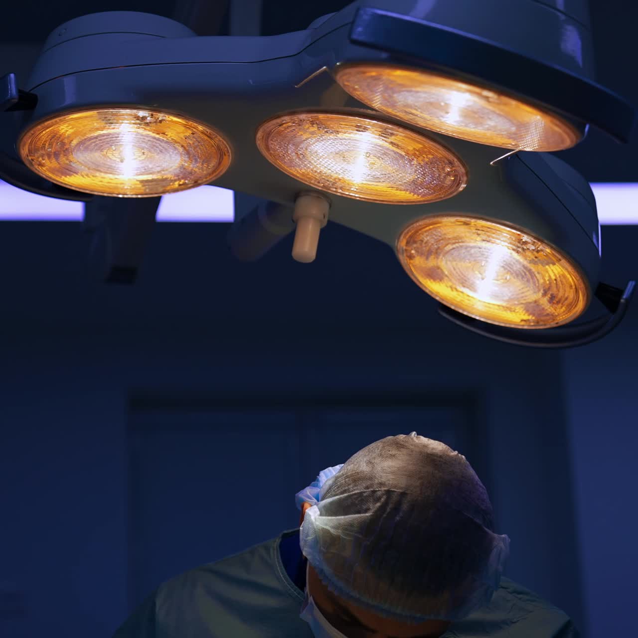 Not bright illumination in the surgical room. Doctor in cap and mask bent over the patient