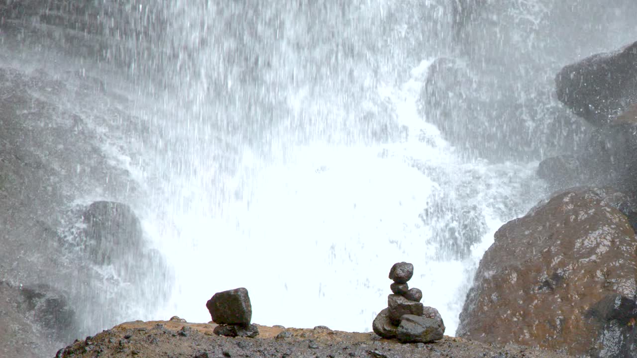 Inquadratura statica di ometti di pietre in equilibrio su massi bagnati davanti a una impetuosa cascata tropicale