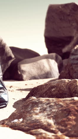 a black garbage bag lays on a sandy beach near large rocks