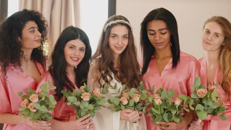 group of multiethnic female friends and bride looking at camera, wearing silk pink and white nighdresses while holding bouquets