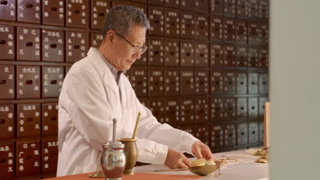 Practicante-De-Retrato-De-Medicina-Tradicional-China-Con-Hierbas-Y-Especias-En-Mortero-Y-Pestil-De-Fondo-De-Madera-Marrón,-Médico-Sosteniendo-Un-Paquete-De-Medicina-Para-Publicidad