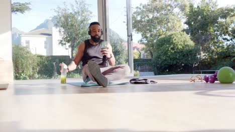 happy african american man resting on mat using smartphone and headphones at sunny home, slow motion