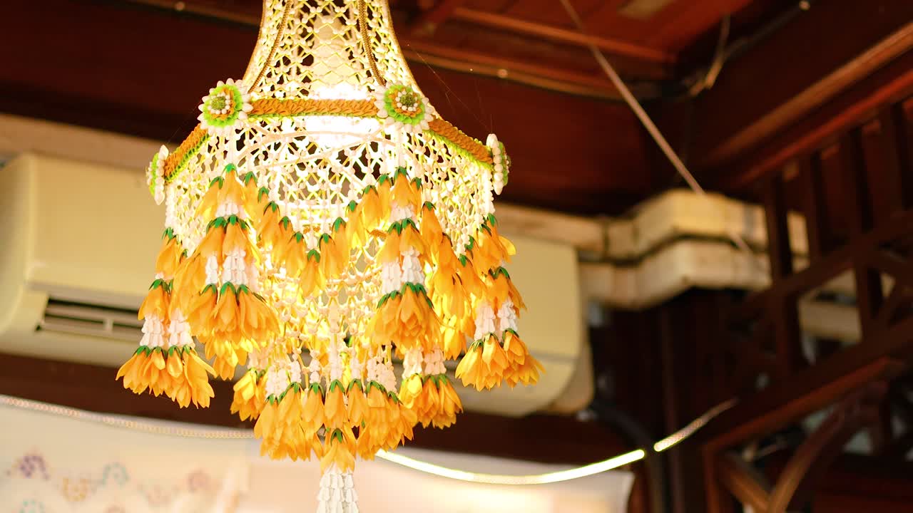 A vibrant Thai flower mobile hangs indoors, illuminated warmly. The intricate design and bright colors create a festive atmosphere