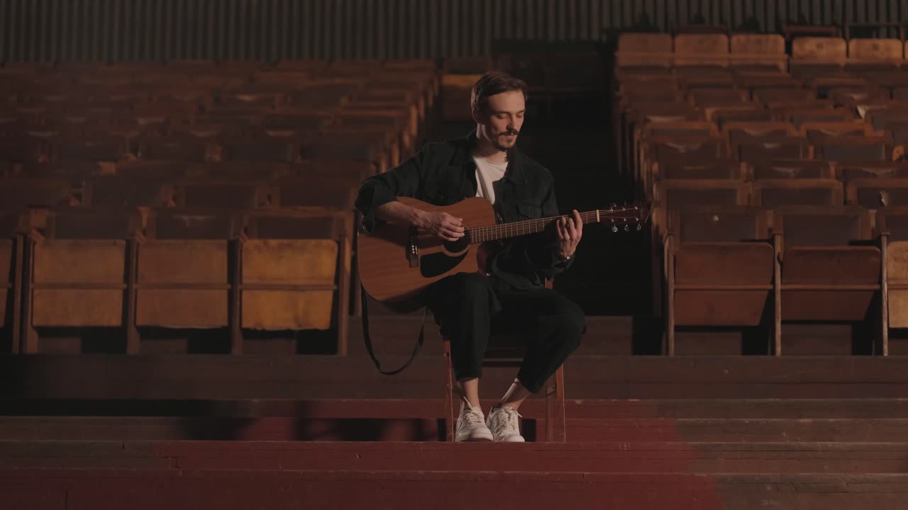 un chico guapo toca una guitarra acústica en un cine abandonado. el músico canta una canción y acompaña en la guitarra
