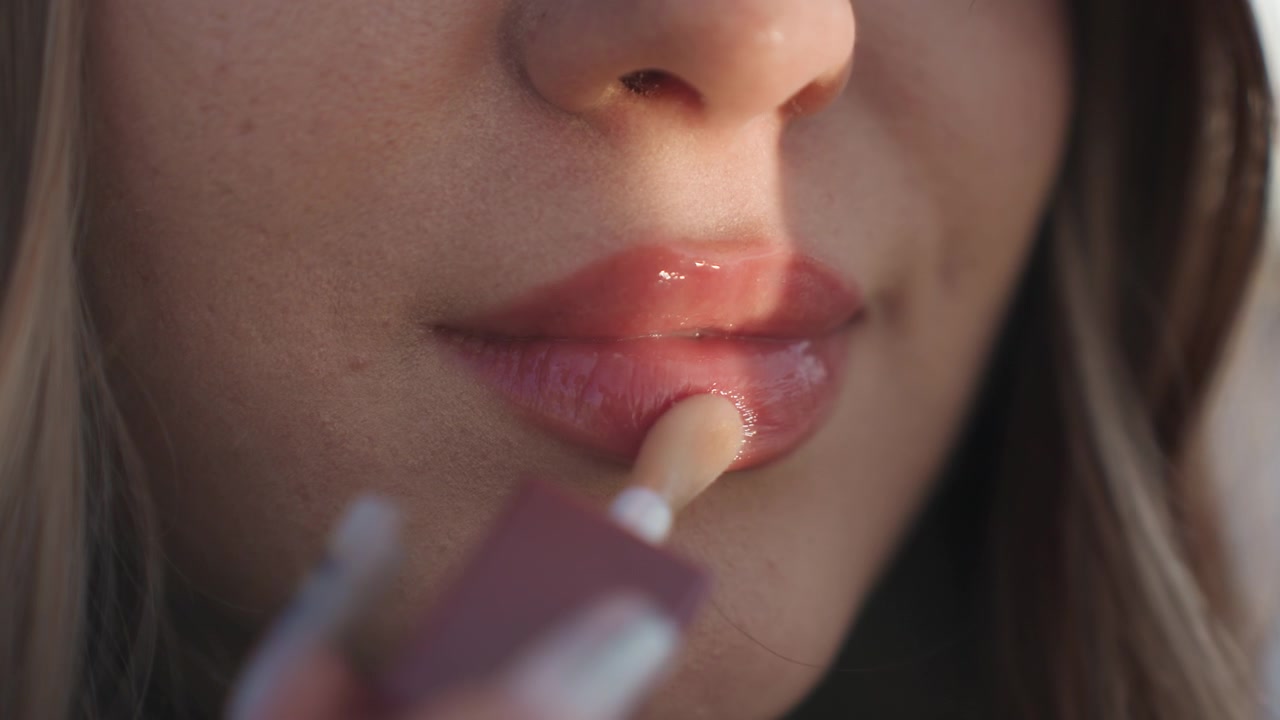 Intimt nærbillede af påføring af blank læbestift, gylden time-glimmer på underlæben, reflekterende glans og glat tekstur, influencer-stemning med diskrete fregner, poleret finish og aften-glamour-stemning