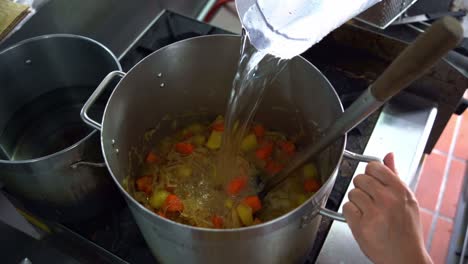 chef pouring a cup of water into a big stock pot full of potatoes, carrots and onions on stovetop, chef cooking in the kitchen, over the shoulder view