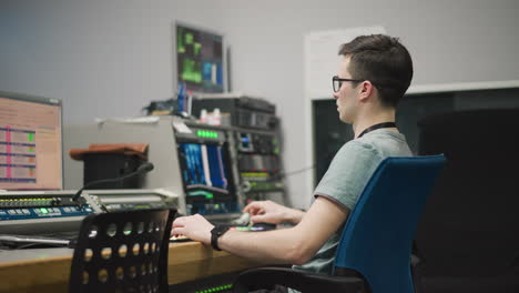 side profile of male video production operator sitting at control panel. focus on hand adjusting equipment with multiple monitors in background. high-tech broadcast studio environment