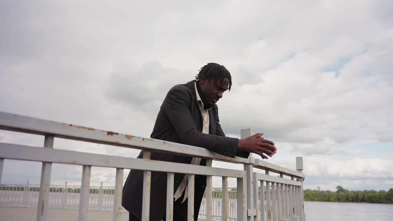 African man walks beside iron rail near calm river under cloudy sky, dressed in coat and shirt, creating thoughtful urban mood with natural light and perspective filled with depth and quiet emotion