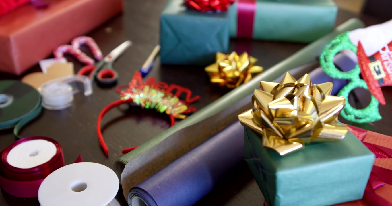 La cámara se desplaza hacia la derecha sobre una mesa, enfocando un regalo verde con un lazo dorado para destacar los regalos terminados.