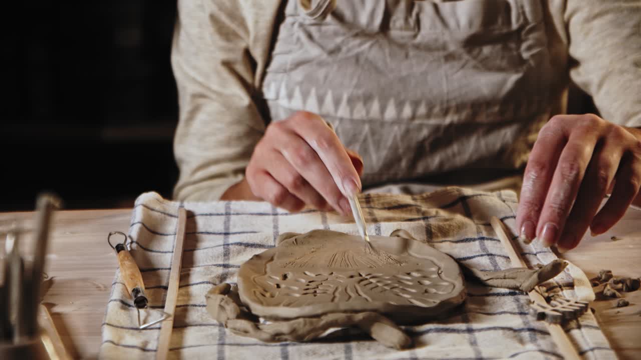 Young woman potter makes patterns on the wet clay plate using a tool