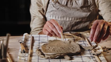 young woman potter makes patterns on the wet clay plate using a tool