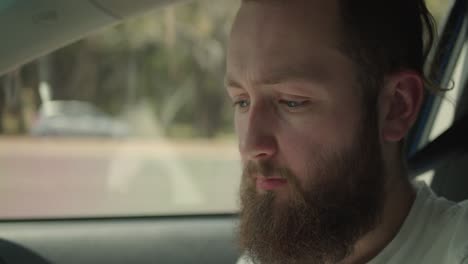 a man with a full beard sits inside a car, his expression shifting between deep thought, curiosity, and slight amusement, framed by soft natural light filtering through the window as he talks