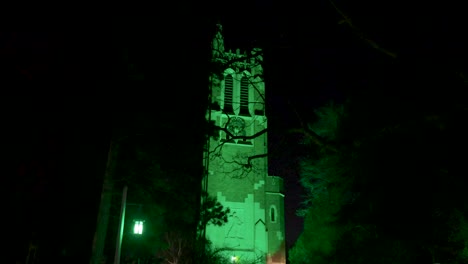 beaumont tower on the campus of michigan state university lit up at night in green in honor of the victims of the february, 2023 mass shooting with gimbal video walking by trees forward