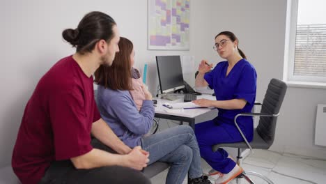 Young-happy-family-a-brunette-guy-in-a-red-T-shirt-together-with-his-wife-and-little-daughter-take-advice-from-a-childrens-doctor-and-a-girl-pediatrician-in-a-blue-uniform-and-glasses-in-a-modern-clinic