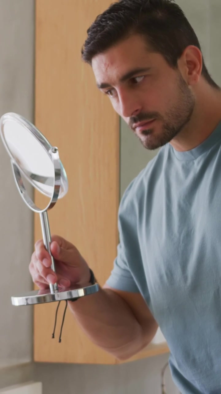Video vertical: Hombre levantando un espejo, inclinándolo en el tocador, inspeccionando la barba incipiente, con reloj y en camiseta.