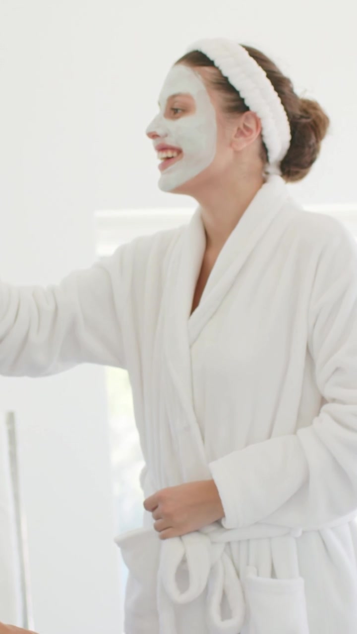 Vertical video: Smiling woman with headband and mask accepting product from left in bath in robe