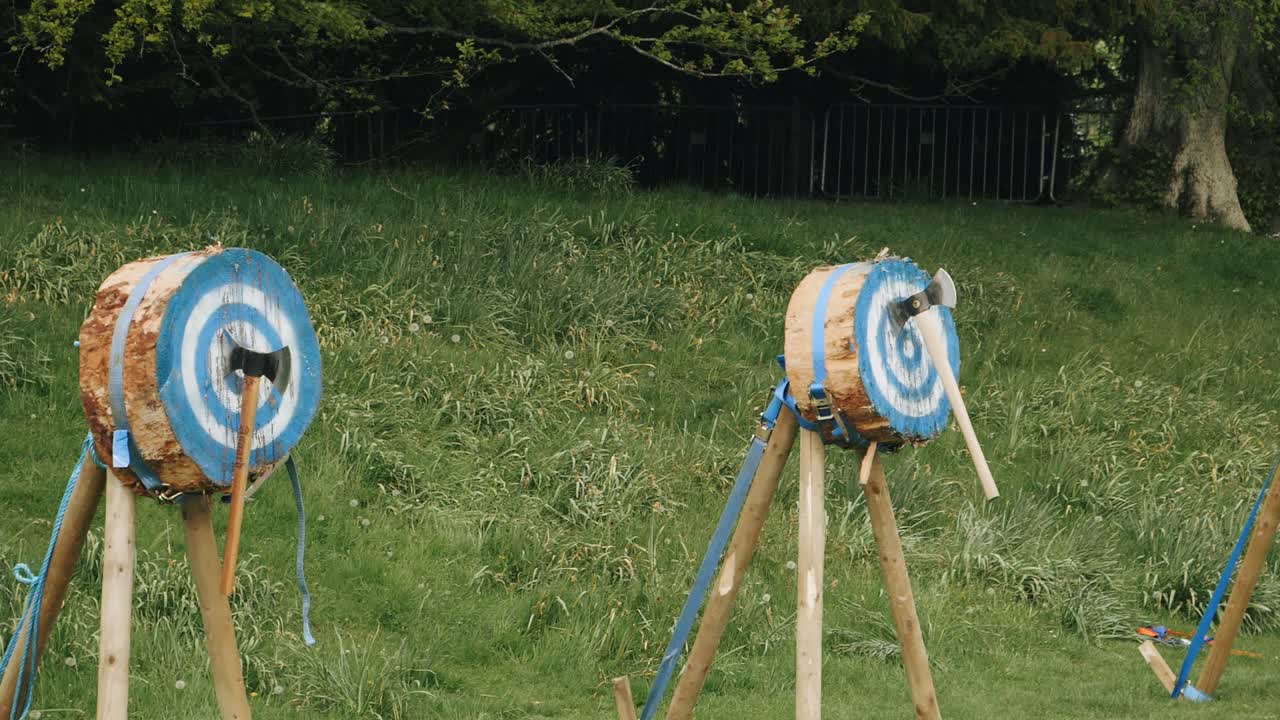 Slow motion footage of traditional axe throwing. Two axes are thrown towards target logs and are embedded. Wide shot showing two empty targets and two axes hitting in succession.