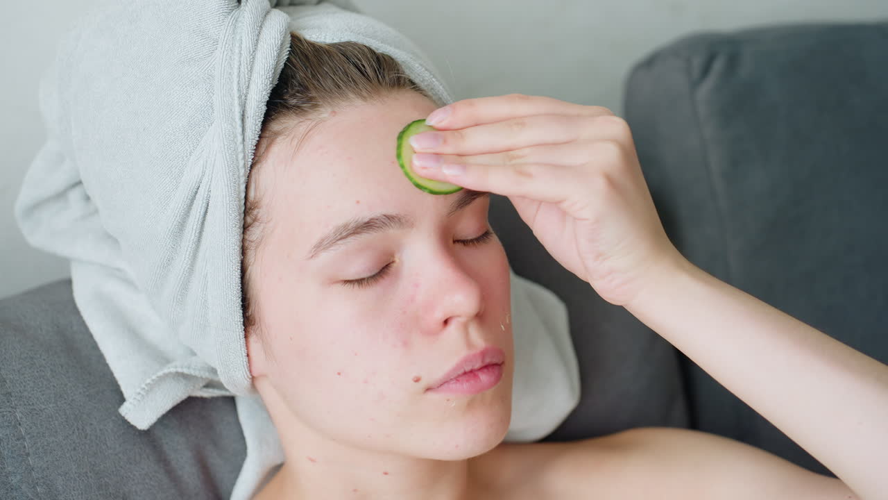 Close-up of woman with towel wrapped around head calmly applying cucumber on forehead as part of refreshing skincare routine, closed eyes and soft lighting creating peaceful, soothing atmosphere