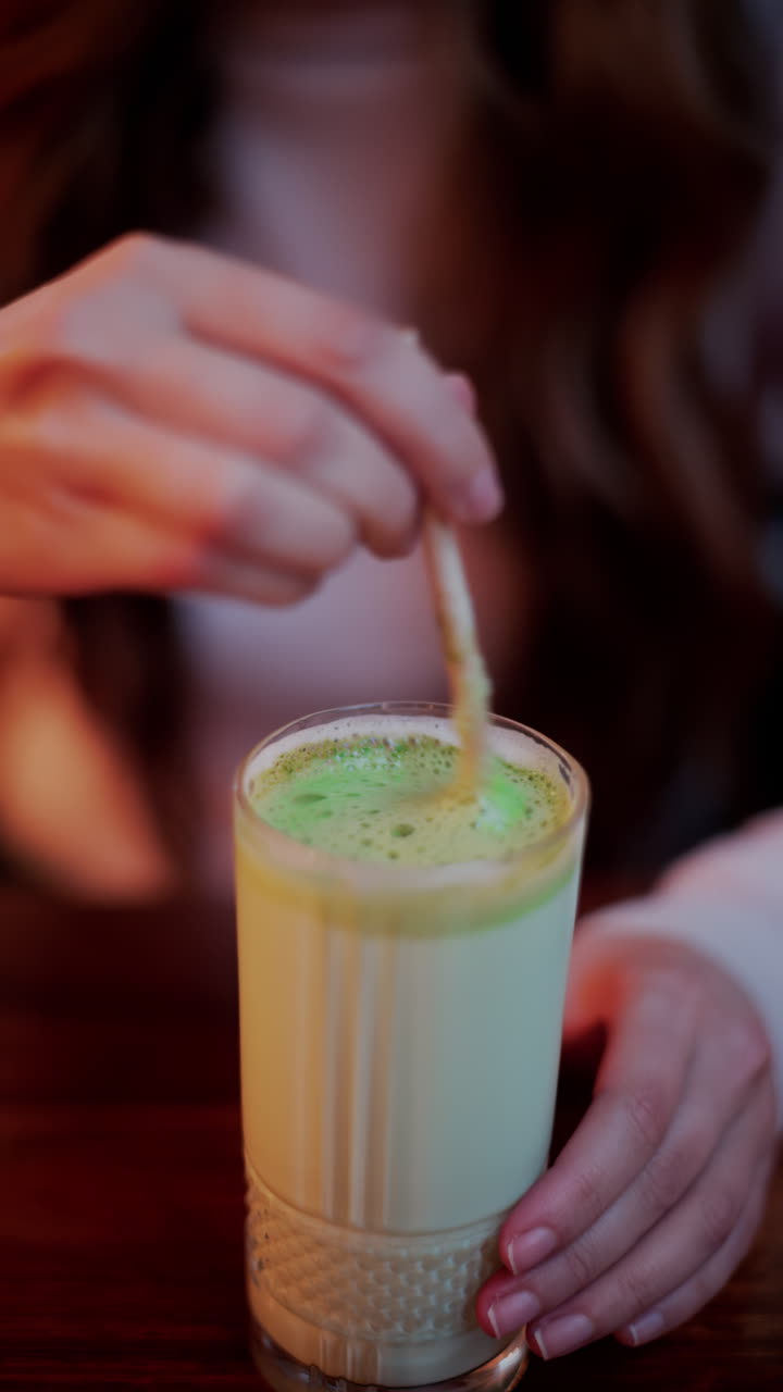 Close up of a woman mixing a matcha latte with a paper straw at a cafe. Vertical
