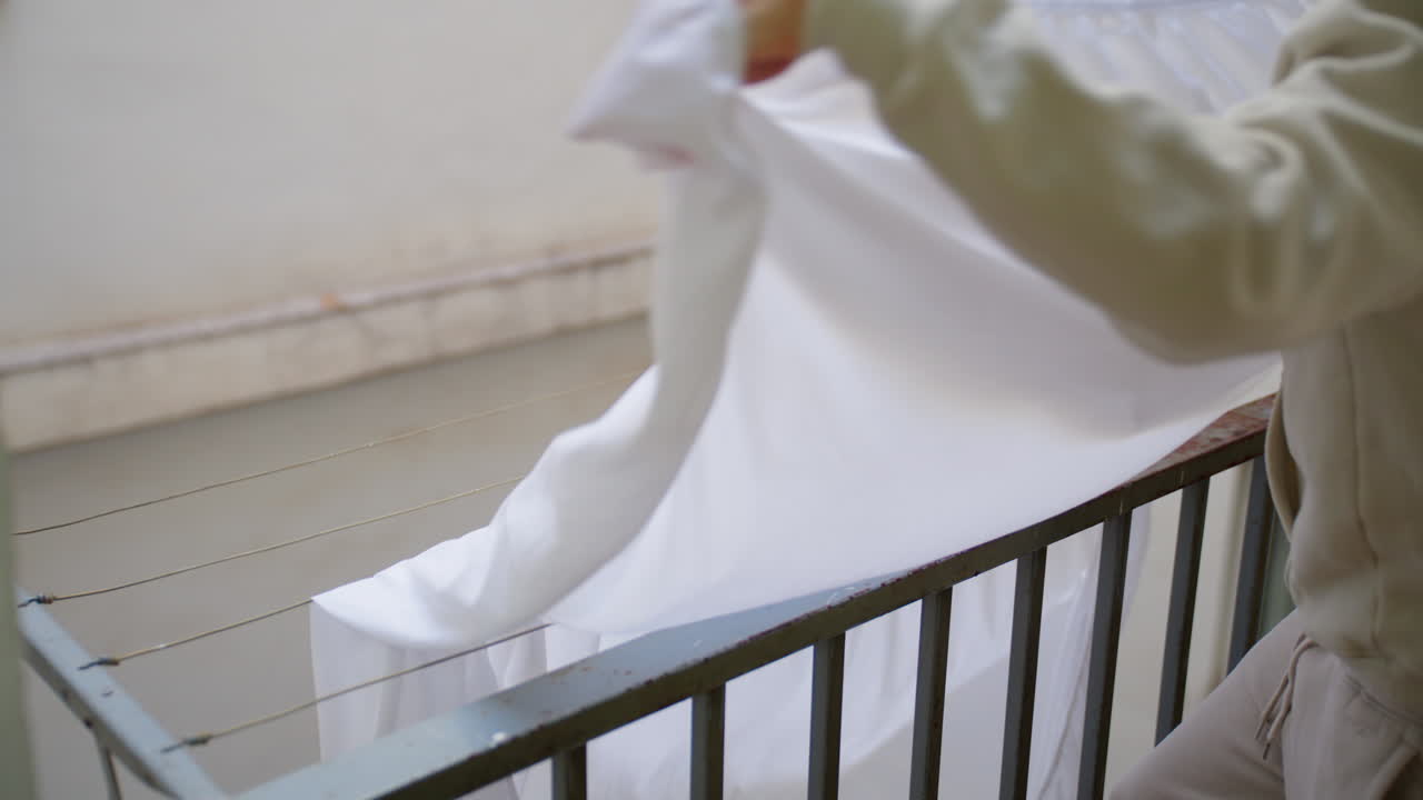 Un giovane fa il bucato sul balcone del suo appartamento. Appende un lenzuolo bianco pulito sullo stendibiancheria, fissandolo con una molletta.