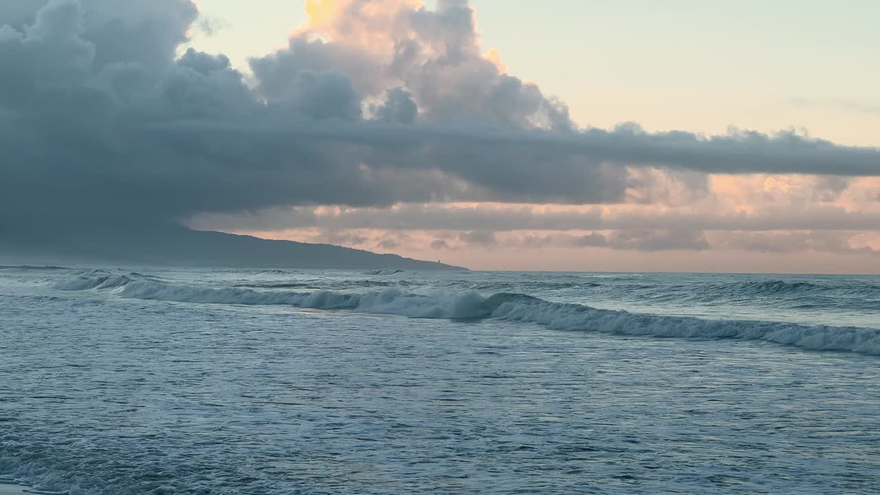 A wide view of a deserted beach with strong waves crashing under a cloudy sunset sky