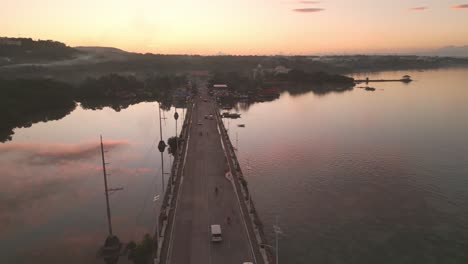 Vista-Aérea-De-La-Hora-Dorada-Del-Tráfico-Del-Puente-Dauis-Y-El-Reflejo-Del-Cielo-Sobre-El-Agua