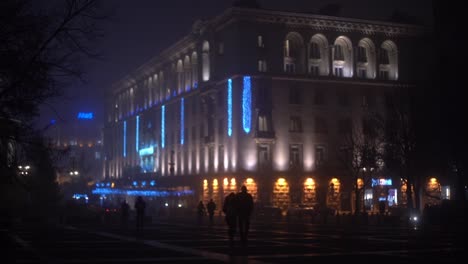 Blue-and-purple-light-up-an-urban-building-as-people-walk-towards-club-at-night