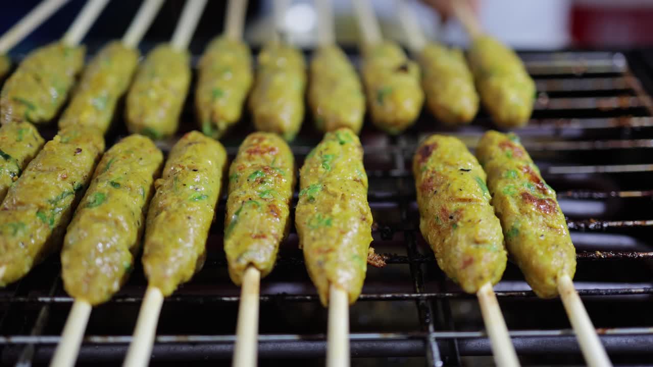 Close-up, panning shot of Thai-styled grilled fish cake with herbs on skewers being cooked on hot flaming charcoal grill by a food vendor