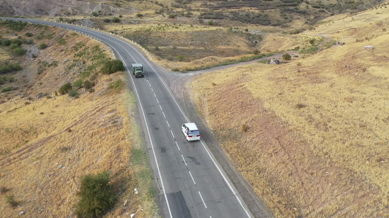 Drone segue l'autostrada che attraversa terreni agricoli aridi e pianure ondulate sotto un cielo sereno, tracciando un camper