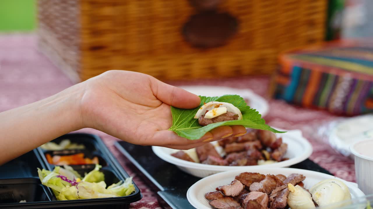 Close-up of a woman's hands making a traditional Korean Ssam by layering grilled beef, garlic, and herbs on a fresh perilla leaf during an outdoor summer picnic