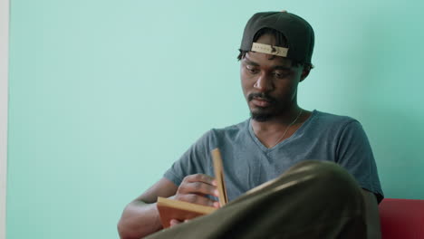 College student in casual wear and cap sits on red couch with crossed leg, opening novel to read while focusing calmly in bright indoor space with turquoise wall