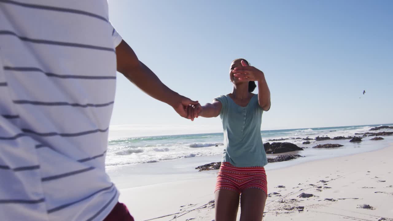 아프리카계 미국인 부부가 미소 짓고 손을 잡고 해변에서 달리고 있습니다.