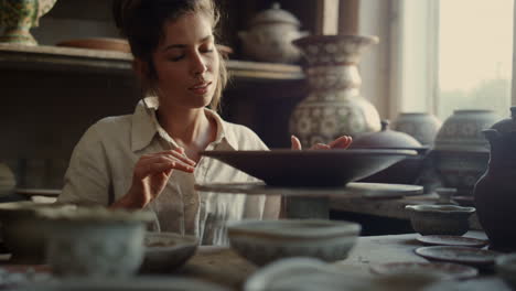 smiling lady touching plate in pottery. artist enjoying clay product in workshop