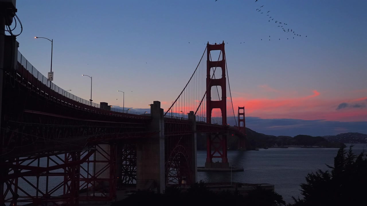 Fugler i V-formasjon over Golden Gate Bridge, San Francisco, California. Fort Point National Historic Site, Battery Vista Point, Velkomstsenter. Oransje, rosa og rød solnedgang over Bay Area og Stillehavet. Statisk bybilde.