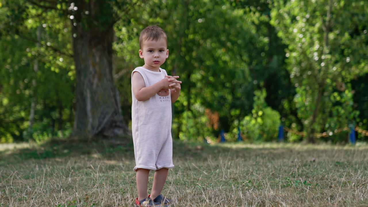 Thoughtful baby boy in a bodysuit stands on the green grass. Calm little toddler looking around outdoors.