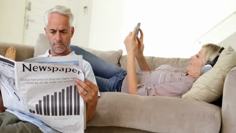 Man-reading-paper-while-partner-is-listening-to-music-on-the-couch