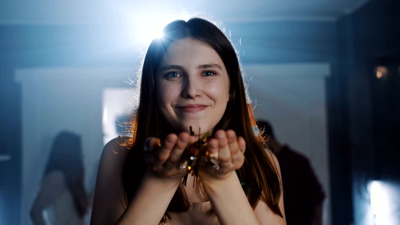 Close-up happy attractive young Caucasian woman blowing out golden confetti and smiling at birthday party slow motion.