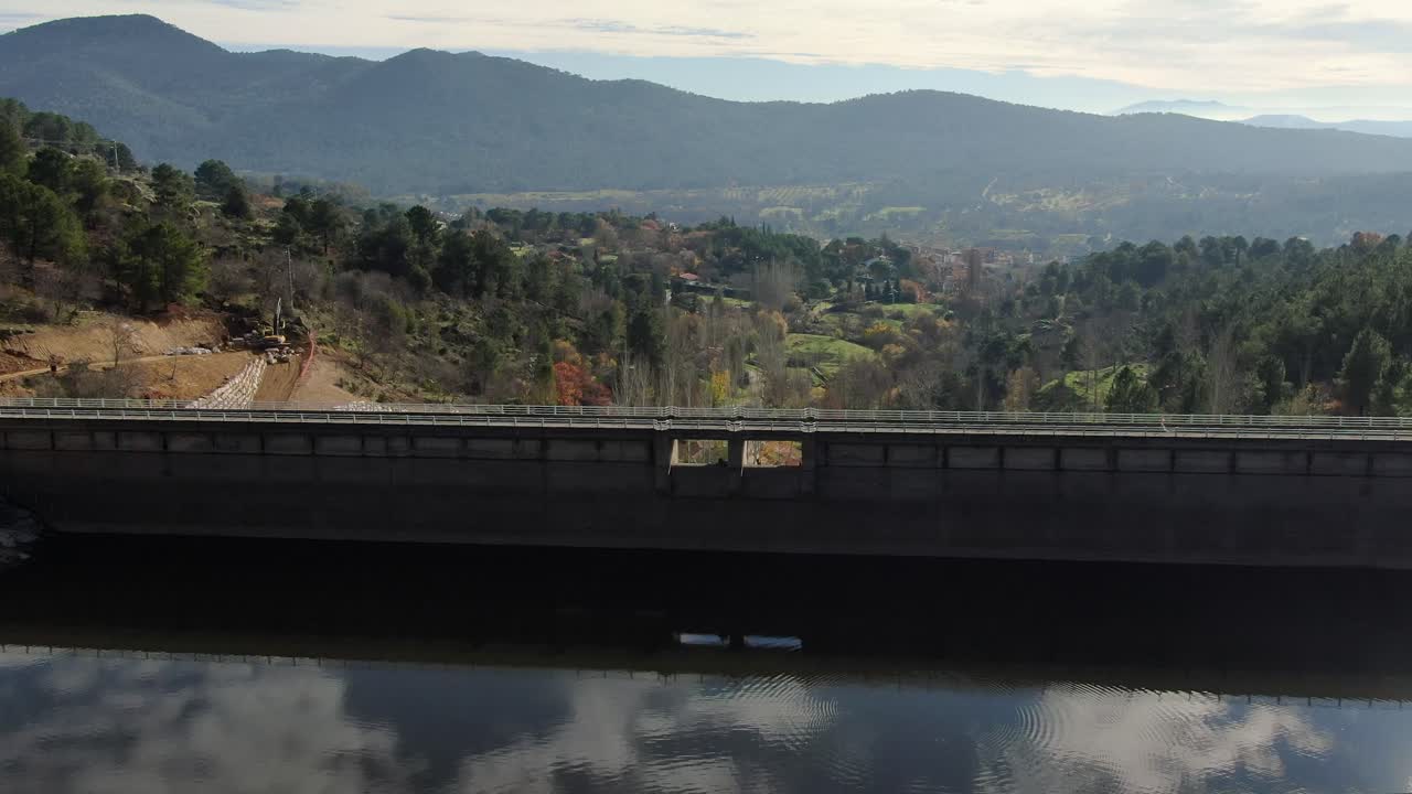 Drone flight over a dam in the Tiétar valley where we see the reflection of the sky in the water and passing the retaining wall we see the work of a road, the valley appears in the background