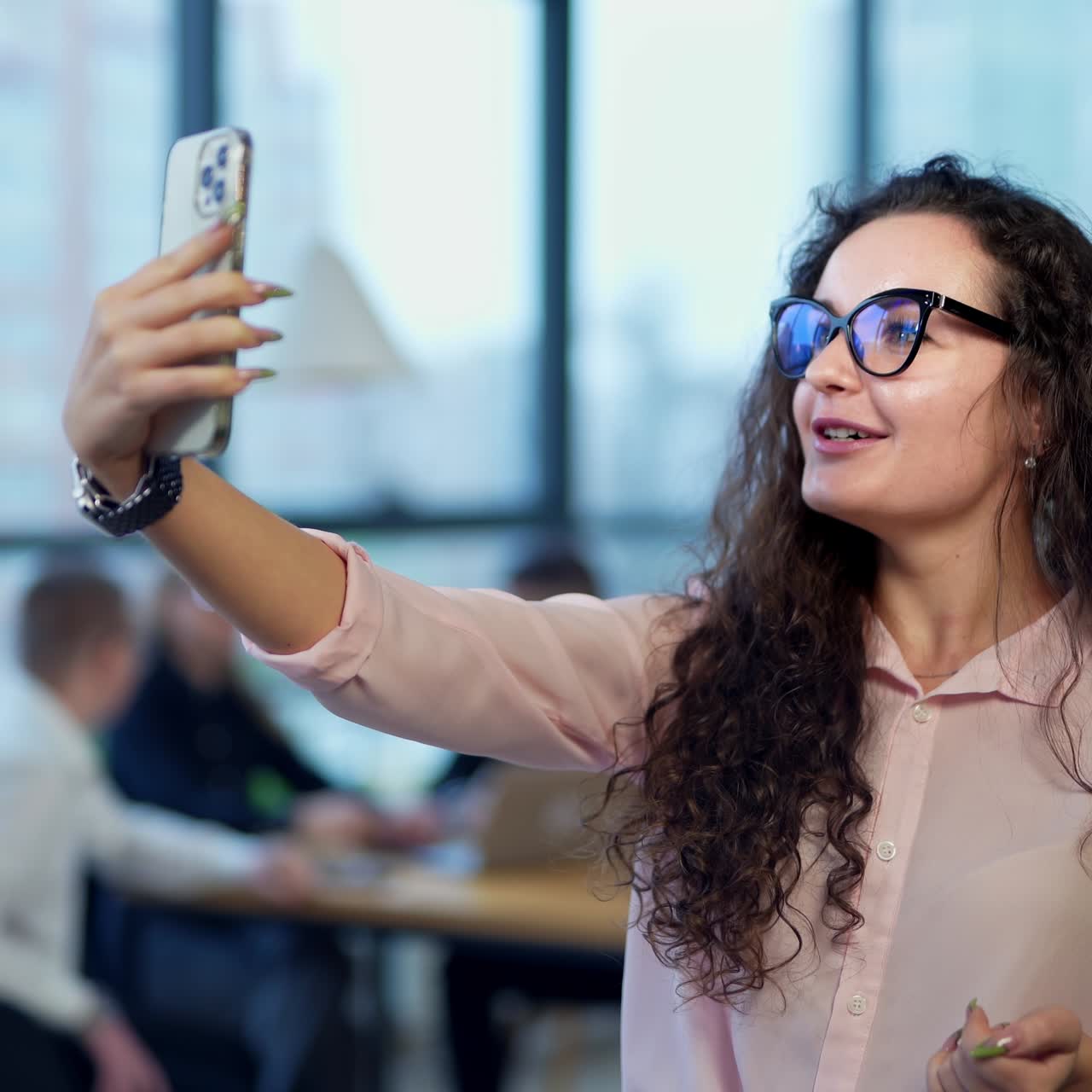 Charming lady with long dark curly hair and glasses takes video on her phone. Office team working at backdrop in blur