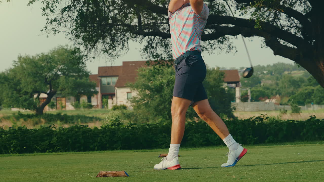 A male golfer drives the ball with force from the tee, filmed from behind at shoulder level. His powerful swing is set against the backdrop of a luxury country club clubhouse in golden light.