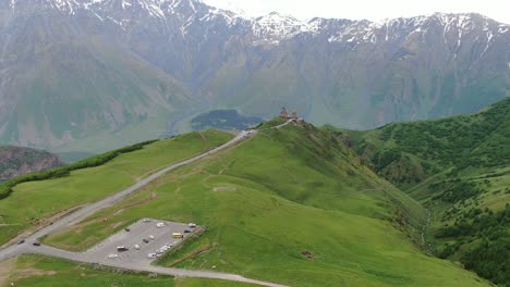 drone aerial view in georgia flying towards gergeti trinity orthodox church in kazbegi surrounded by green mountains valley with snowed peaks