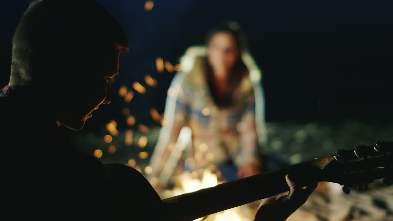 그의 여자 친구에게 기타를 연주하는 남자는 저녁에 모닥불 주위에 서로 옆에 앉아